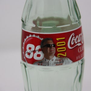May include: A clear glass Coca-Cola bottle with a red label featuring a photo of Dale Jarrett and the text "88 2001 Coca-Cola Classic 8 FL OZ".