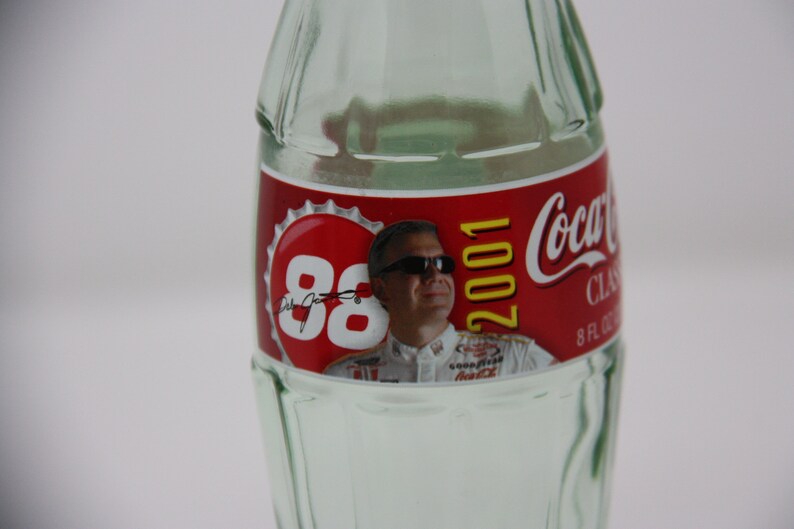 May include: A clear glass Coca-Cola bottle with a red label featuring a photo of Dale Jarrett and the text "88 2001 Coca-Cola Classic 8 FL OZ".