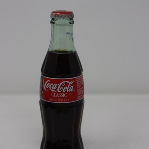 May include: A glass bottle of Coca-Cola Classic, 8 fl oz (237 ml), with a red label and a green cap.
