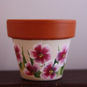 Op de afbeelding: Een terracotta bloempot met een witte basis beschilderd met roze bloemen en groene bladeren.