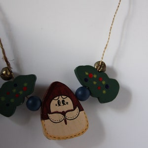 May include: A wooden necklace featuring a green, red and yellow painted tree, a brown and beige painted figure, and blue beads. The necklace is strung on brown twine.