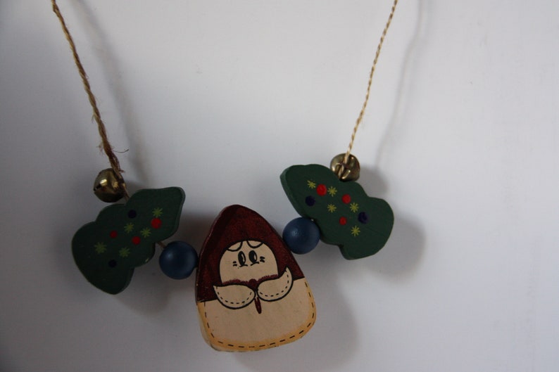 May include: A wooden necklace featuring a green, red and yellow painted tree, a brown and beige painted figure, and blue beads. The necklace is strung on brown twine.
