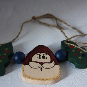 May include: A wooden Christmas garland featuring a girl, two Christmas trees, and blue beads. The girl is wearing a red hat and a white dress. The trees are green with red and yellow decorations.