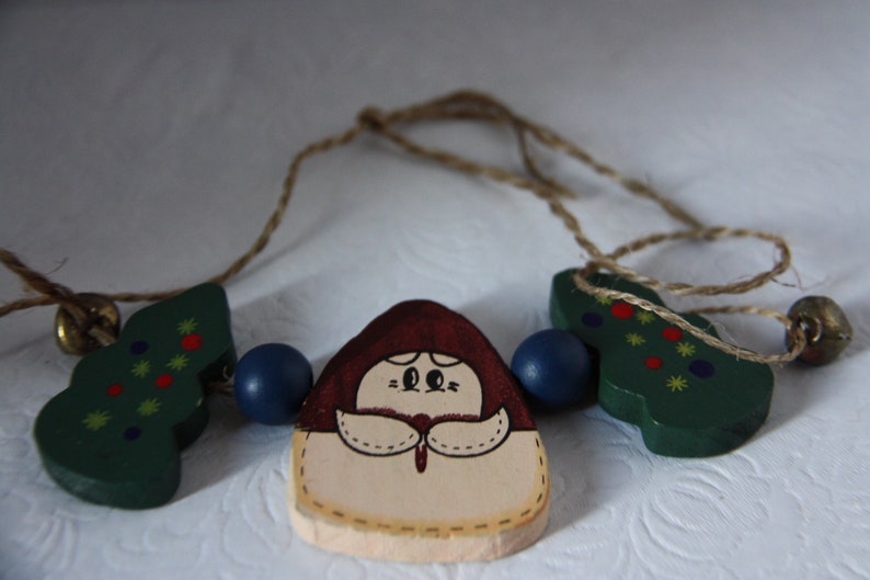 May include: A wooden Christmas garland featuring a girl, two Christmas trees, and blue beads. The girl is wearing a red hat and a white dress. The trees are green with red and yellow decorations.