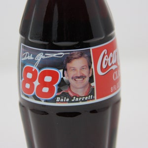 May include: A Coca-Cola Classic 8 fl oz glass bottle with a red, white, and blue label featuring Dale Jarrett's signature and the number 88. The label also includes a photo of Dale Jarrett in his racing uniform.