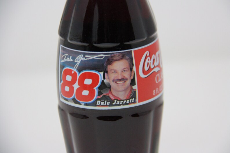 May include: A Coca-Cola Classic 8 fl oz glass bottle with a red, white, and blue label featuring Dale Jarrett's signature and the number 88. The label also includes a photo of Dale Jarrett in his racing uniform.