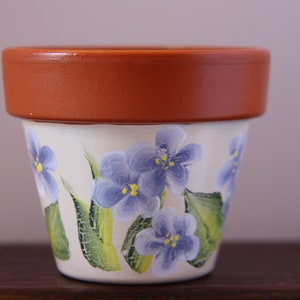 Op de afbeelding: Bloempot met een terracotta rand en een crèmekleurige body. De body is versierd met handgeschilderde blauwe viooltjesbloemen en groene bladeren. Ideaal voor het planten van bloemen en kruiden. Afmetingen in centimeters.