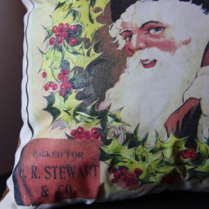 May include: A white pillow with a vintage illustration of Santa Claus holding a bag of gifts. The illustration is surrounded by holly leaves and red berries. The pillow has a red label that says "Packed for E.R. Stewart & Co."