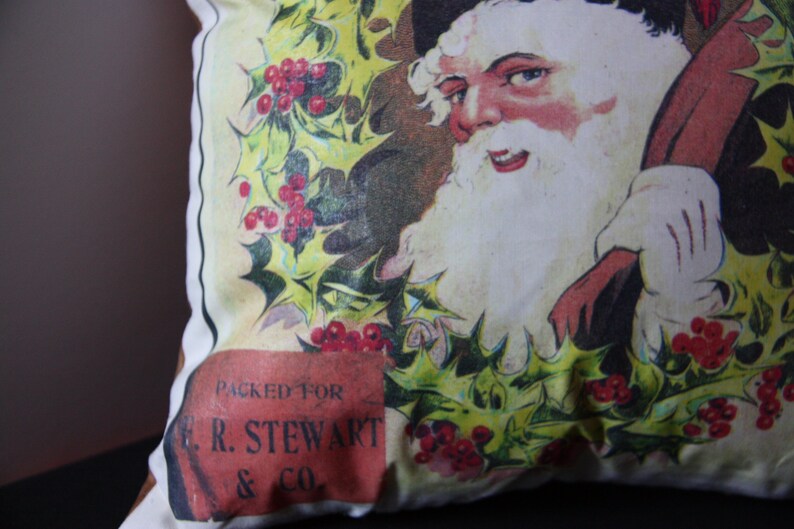 May include: A white pillow with a vintage illustration of Santa Claus holding a bag of gifts. The illustration is surrounded by holly leaves and red berries. The pillow has a red label that says "Packed for E.R. Stewart & Co."