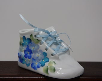 May include: A white ceramic baby shoe with blue and yellow floral designs and a light blue satin ribbon tied around the laces.
