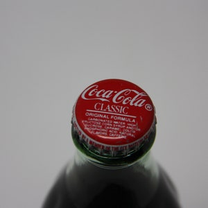 May include: A red and white Coca-Cola Classic bottle cap with the text "Coca-Cola Classic Original Formula Carbonated Water High Fructose Corn Syrup And/Or Sucrose Caramel Color Phosphoric Acid Natural Flavors Caffeine"