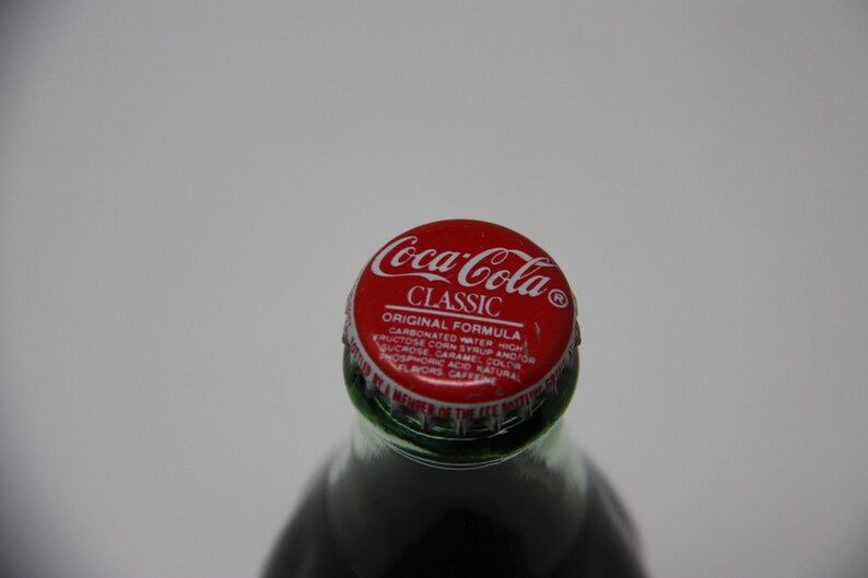 May include: A red and white Coca-Cola Classic bottle cap with the text "Coca-Cola Classic Original Formula Carbonated Water High Fructose Corn Syrup And/Or Sucrose Caramel Color Phosphoric Acid Natural Flavors Caffeine"