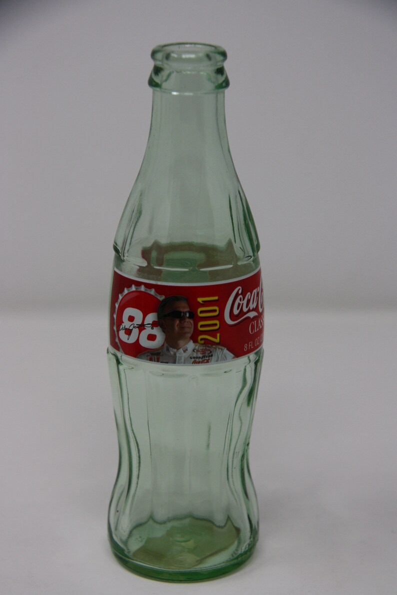 May include: A clear glass Coca-Cola bottle with a red label featuring a photo of a race car driver and the text "88 2001 Coca-Cola Classic 8 fl oz".