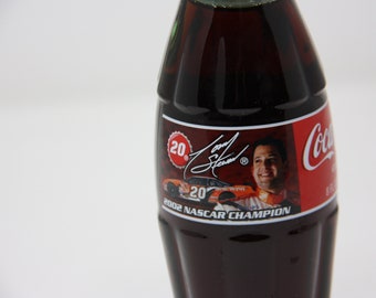 May include: A Coca-Cola bottle with a red, white, and black label featuring a photo of a race car driver, his signature, and the text "20. 2002 NASCAR CHAMPION".