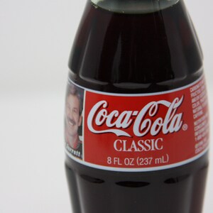 May include: A glass bottle of Coca-Cola Classic soda. The bottle is dark brown and has a red label with the Coca-Cola logo and the words "Classic" and "8 fl oz (237 ml)".