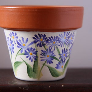Op de afbeelding: Een terracotta bloempot met een witte achtergrond en handgeschilderde paarse bloemen met gele centra.