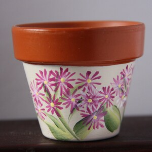 Pode incluir: Um vaso de terracota com base branca pintada com flores cor-de-rosa e folhas verdes.