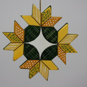 May include: A handcrafted, star-shaped ornament with a geometric design. The ornament features yellow, green, and white painted patterns, including stripes, polka dots, and plaid. It is suspended by a thin, gold-colored string, perfect for holiday decor.