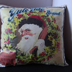 May include: A white pillow with a vintage illustration of Santa Claus holding a bag of gifts. The illustration is surrounded by a wreath of holly and berries. The text "Yule Tide Brand" is printed in blue and red letters at the top of the pillow. The text "Packed for E.R. Stewart & Co." is printed at the bottom of the pillow.