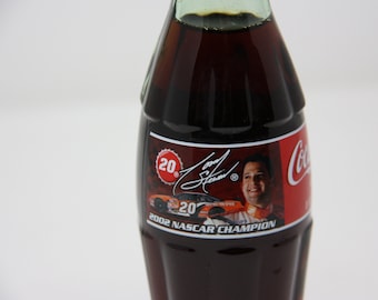 May include: A Coca-Cola bottle with a red, white, and black label featuring a photo of a race car driver and the text "2002 NASCAR CHAMPION".