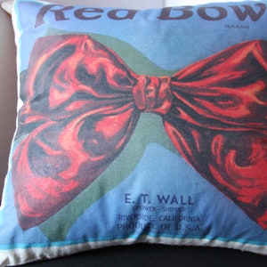 K&ouml;nnte beinhalten: Ein wei&szlig;es Kissen mit einer roten Schleife, die darauf gedruckt ist. Der Text "Rea BOW BRAND" ist oben auf dem Kissen gedruckt. Der Text "E. T. WALL GROWER-SHIPPER RIVERSIDE, CALIFORNIA PRODUCE OF U.S.A" ist unten auf dem Kissen gedruckt.