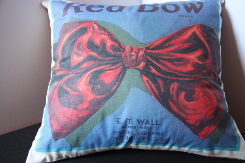 K&ouml;nnte beinhalten: Ein wei&szlig;es Kissen mit einer roten Schleife, die darauf gedruckt ist. Der Text "Rea BOW BRAND" ist oben auf dem Kissen gedruckt. Der Text "E. T. WALL GROWER-SHIPPER RIVERSIDE, CALIFORNIA PRODUCE OF U.S.A" ist unten auf dem Kissen gedruckt.