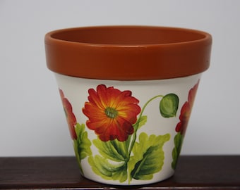 May include: A terracotta flower pot with a white base painted with orange and red flowers and green leaves.