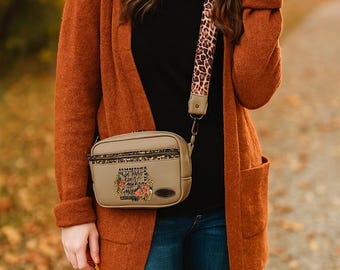 May include: A tan crossbody bag with a leopard print strap. The bag has a floral design and text on the front. The person is wearing a rust-colored cardigan, a black shirt, and blue jeans.