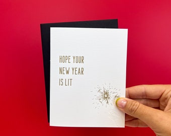 May include: White card with gold lettering that reads "Hope your new year is lit" with a gold glitter design of a firework.