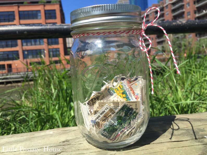 May include: A clear glass jar with a silver lid and a red and white twine bow. The jar is filled with vintage postage stamps.