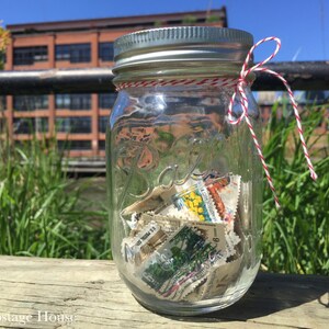 May include: A glass jar filled with colorful postage stamps, tied with a red and white twine bow. The jar is sitting on a wooden surface with green grass and a brick building in the background.