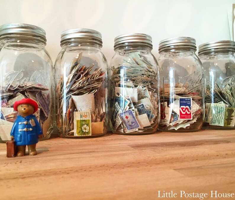 May include: Five glass jars filled with colorful postage stamps. The jars are lined up on a wooden surface. A small blue teddy bear with a red hat is in the foreground.