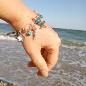 Op de afbeelding: Een zilveren armband met blauwe en oranje kralen, een zeester en schelpbedels. De armband wordt gedragen op de pols van een persoon tegen een achtergrond van blauw water en wit zand.