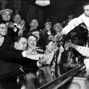 May include: A black and white photo of a group of people gathered around a bar, some are holding glasses and drinking. The people are dressed in 1940s clothing. The bar is made of dark wood and has a polished surface. There are bottles and glasses on the bar. The people are smiling and laughing. The photo is taken in a dimly lit room. The photo is a vintage photo.