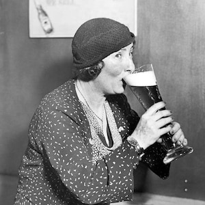 May include: A person wearing a black and white patterned dress and a black hat is drinking from a tall glass of beer.