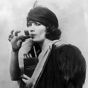 May include: A black and white photo of a woman smoking a cigarette holder. She is wearing a dark-colored turban and a dark coat. She is holding a small, dark-colored case that says 