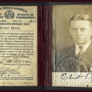 May include: A black and white photo of Eliot Ness, a man with dark hair, wearing a suit and tie. The photo is on a white card with the text 