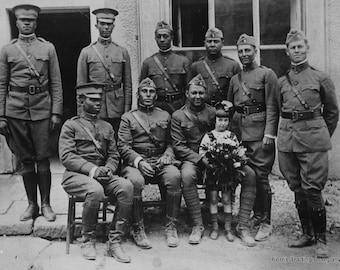 May include: A group of men in military uniforms stand and sit in front of a building. A young girl in a dress holds a bouquet of flowers. The men are all African American and are wearing the same uniform. The girl is standing in front of the men and is smiling. The building is a brick building with a door and windows. The photo is in black and white.