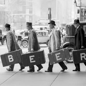 May include: Four men in black and white clothing are walking down a city street carrying suitcases. The suitcases are black and have white letters on them that spell out 