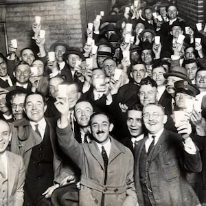 May include: A black and white photo of a group of people in a hallway, all raising their glasses in a toast. The people are dressed in suits and ties, and they are all smiling. The photo is taken from a low angle, and the people are all looking up at the camera.