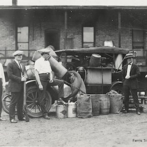 May include: A black and white photo of five men standing in front of a truck. The truck is loaded with various items, including barrels and cans. The men are all wearing hats and suits. The truck is parked in front of a brick building. The text 