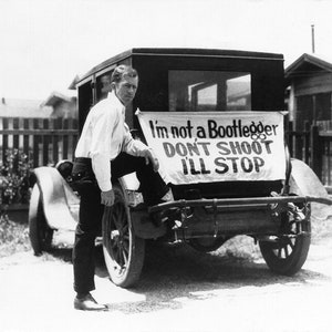 May include: A black and white photo of a man standing next to a car. The car has a sign on the back that says 