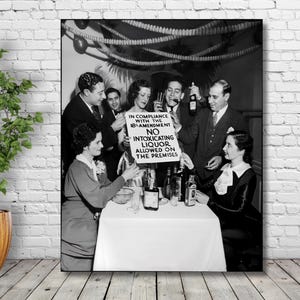 May include: A black and white photograph shows a group of people gathered around a table at a party.  A sign reads: 