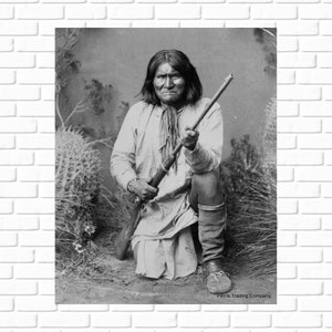 May include: A black and white photograph of a Native American man holding a rifle. He is wearing traditional clothing and is kneeling in a desert landscape. The photo is captioned "Ferns Trading Company."
