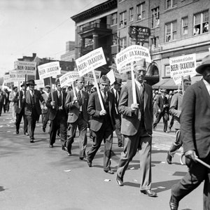 May include: A black and white photo of a group of men marching in a street. The men are holding signs that read 
