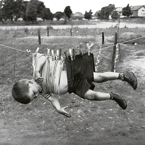 May include: A young boy is hanging upside down from a clothesline with clothespins holding his shirt and shorts to the line. He is wearing a striped shirt and dark shorts. The boy is smiling and appears to be having fun.