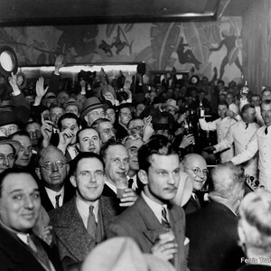 May include: A black and white photo of a crowd of people gathered in a bar. The people are all dressed in 1930s clothing and are looking at something off camera. There are several bartenders in white shirts and black ties serving drinks. The bar has a mural on the wall with a tropical theme. The text 