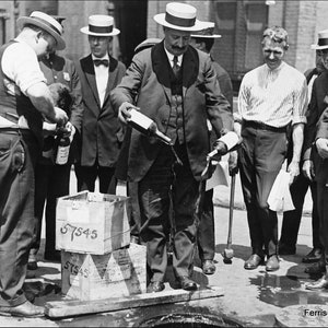 May include: A group of men in suits and hats stand around a wooden platform. One man pours a bottle of liquid onto the platform. The platform has two boxes stacked on top of each other. The boxes are labeled with the number 