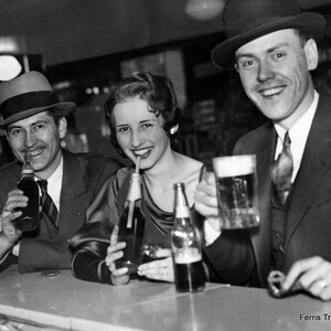 May include: Three people are sitting at a bar counter. The person on the left is wearing a hat and drinking from a bottle. The person in the middle is wearing a dress and drinking from a bottle. The person on the right is wearing a hat and drinking from a glass. The bar counter is made of wood and has a light brown color. The background is a blurry image of a bar with people and lights.