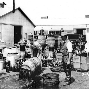 May include: Black and white photo of a group of people working in a warehouse. There are several large metal containers, barrels, and bottles. The photo is dated 3-31-32.